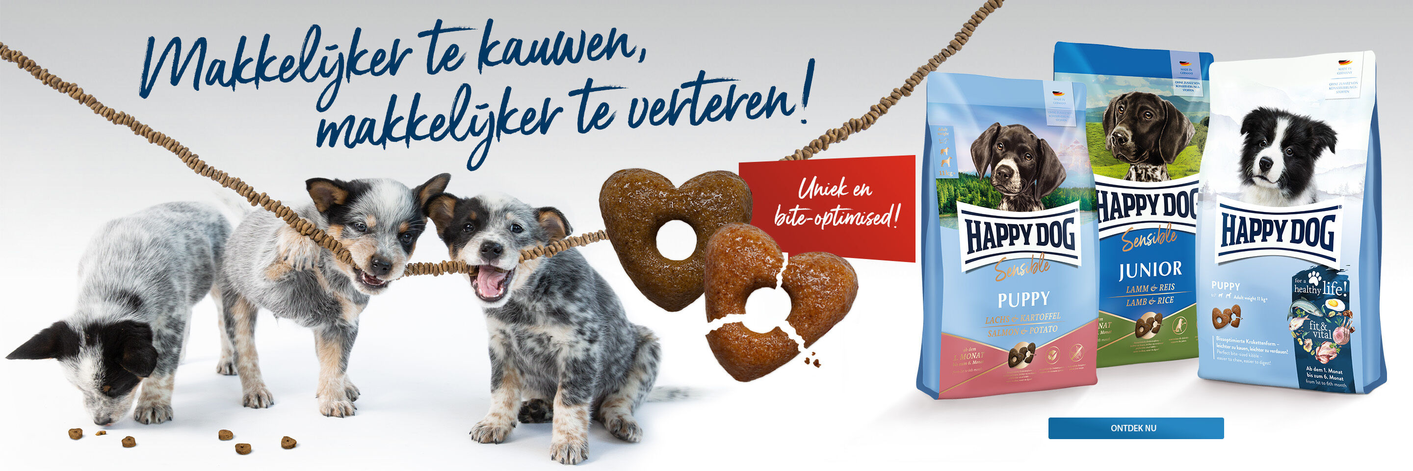 Drei Welpen kauen an einer Schnur mit Kroketten. Daneben stehen Futtersäcke von Happy Dog.