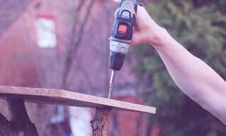 Ein Mann bohrt mit einer Bohrmaschine eine Holzplatte an einem Ast fest.