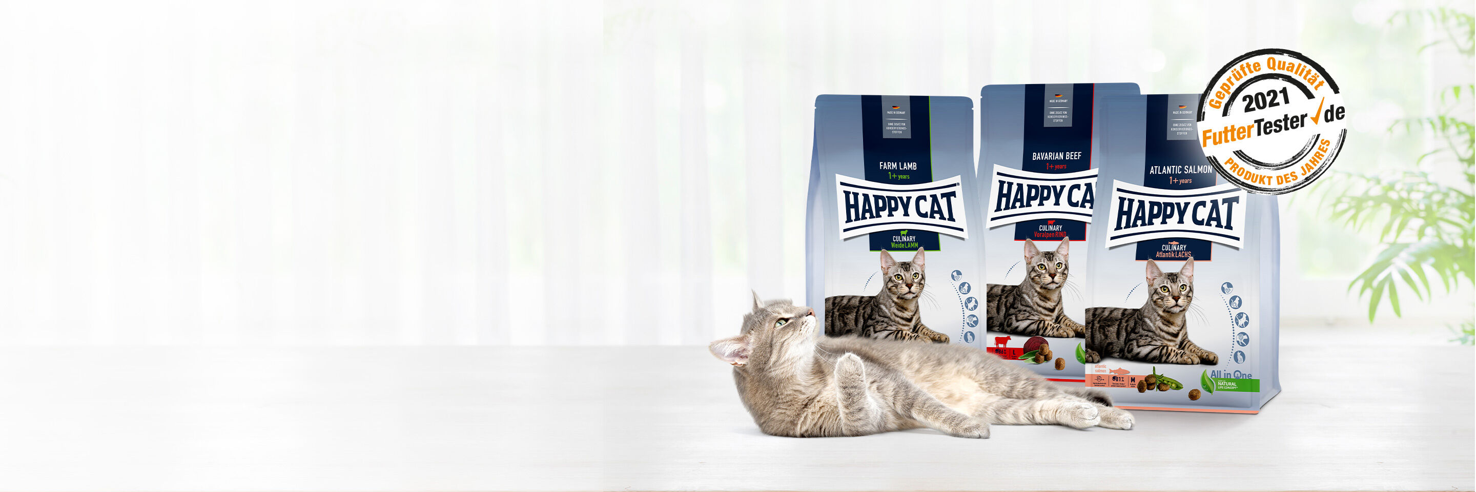 Katze liegt entspannt am Boden vor drei Futterpackungen Happy Cat Trockenfutter 