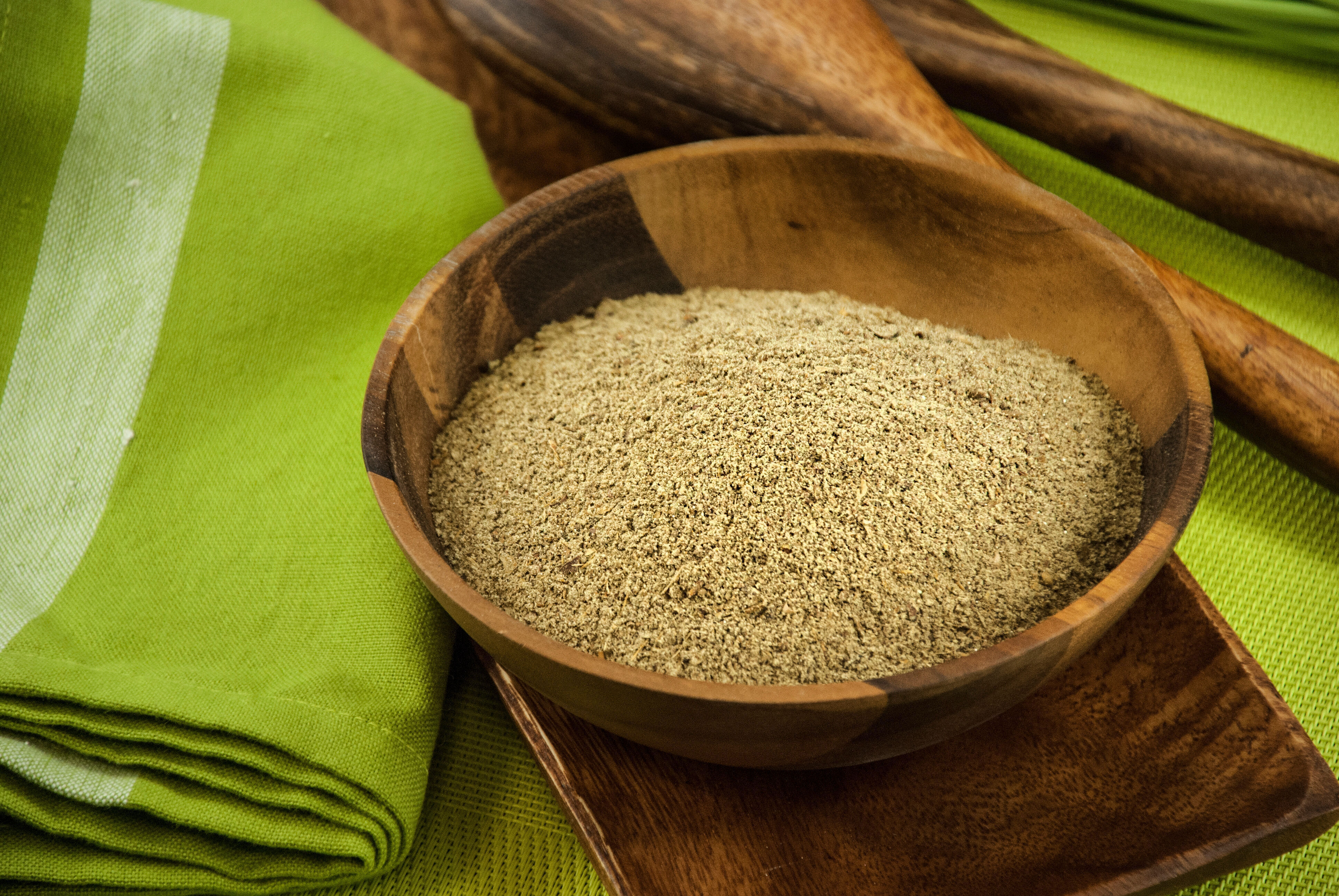 Holzschale voller gemahlenem Gewürz auf einem grünen Tuch, umgeben von Holzlöffeln.
