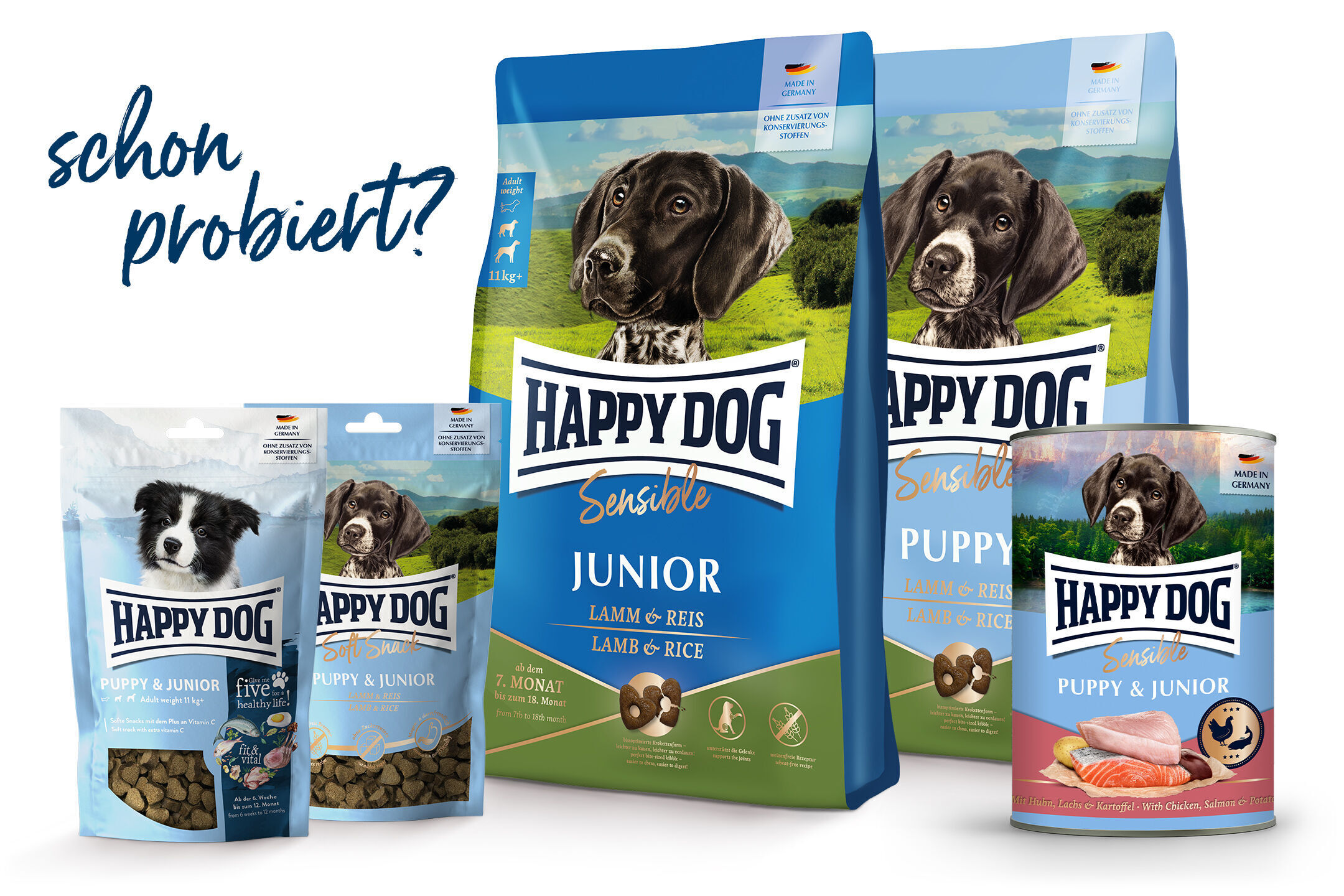 Verpackungen von Happydog-Hundefutter mit verschiedenen Sorten und Größen. Ein großer Beutel in der Mitte zeigt einen Hund auf einer Wiese. Text: "schon probiert?"