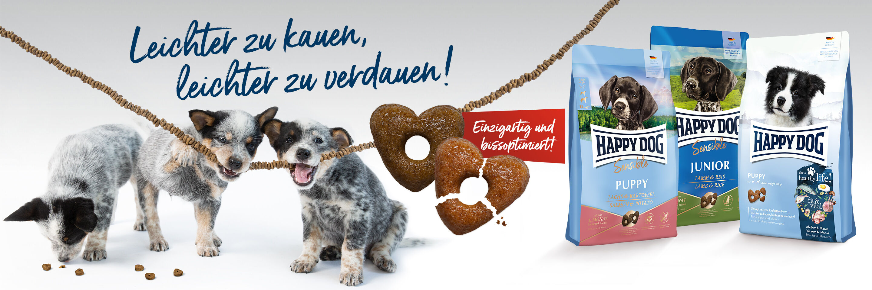 Das Happy Dog Young Trockenfutter wird in Herzkrokettenform gefertigt. Drei Welpen fressen Herzkroketten, die an einer Schnur aufgefädelt wurden.