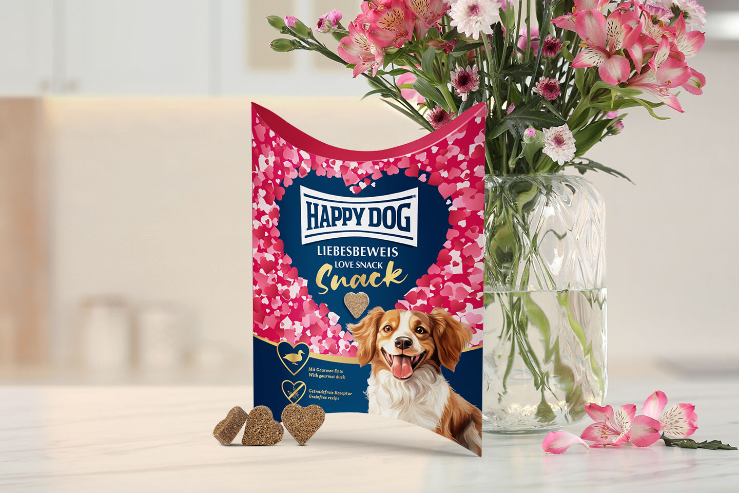 Eine Packung des Happy Dog Valentinssnack "Liebesbeweis" mit einem lächelnden Hund und einer großen Anzahl an angeordneten Herzen steht auf einem Küchentisch vor einem Blumenstrauß in einer Vase.