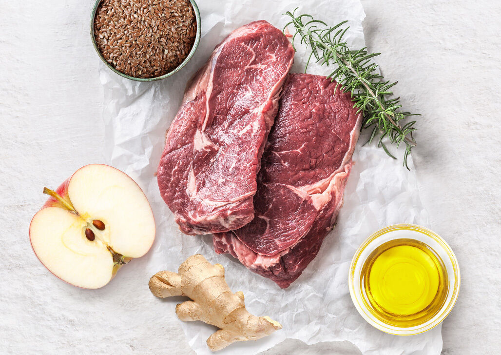 Zwei Scheiben rohes Fleisch, Ingwer, Rosmarin, Apfel, Leinsamen und ein kleines Schälchen Öl