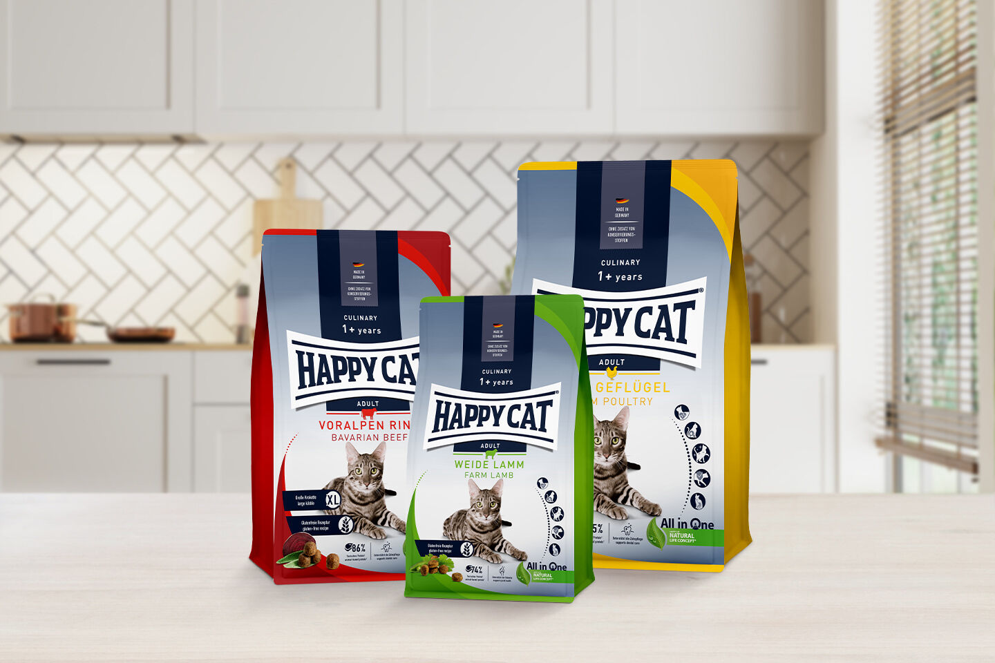 Drei Packungen Happy Cat Katzenfutter für sterilisierte Katzen stehen auf einem Tisch vor einer hellen Küche in unscharfen Hintergrund. Jede Packung zeigt verschiedene Geschmacksrichtungen und besteht aus all natürlichen Zutaten.